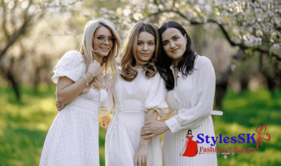 Three women in white dresses smiling together in a blooming orchard